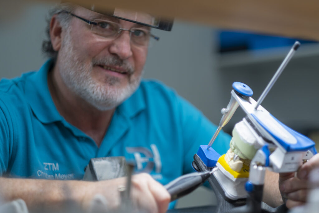 Zahntechniker sitzt an seinem Arbeitsplatz im Labor und lächelt in die Kamera.